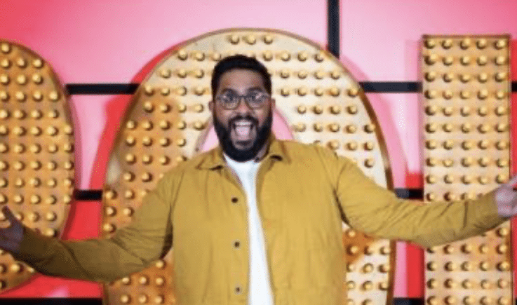 Comedian Eshaan Akbar, wearing a yellow jacket and white tshirt, stands in front of a red and gold Live At The Apollo background with his arms outstretched